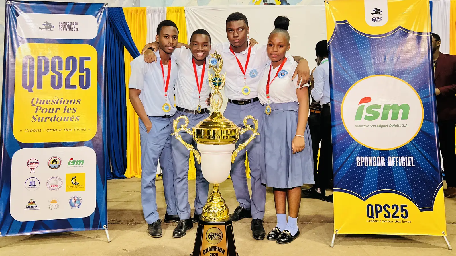 Les quatre champions de QPS25, médaille au cou, posent avec le trophée entre la bannière des partenaires et celle d'ISM, sponsor officiel.