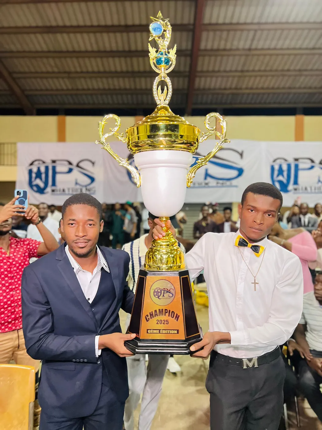 Le trophée doré de champion 2025 est brandi à deux, devant les banderoles du concours.